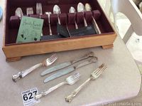 Wooden storage box with burgundy felt slots holding 12 silver plated knives with decorative handles, plus assorted forks and spoons on a table.