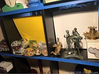 Shelf with various glass paperweights, Alvar Aalto collection bowl box, and metal vase holder with three figures including a mermaid and Poseidon candlestick holder