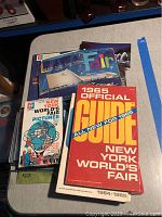 Photo showing a red-covered 1965 Official Guide Book, the Make-A-Model Book, and a flashcard set titled 'New York World's Fair Pictures' on a table.
