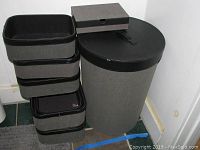 Full set of six grey rectangular storage baskets stacked, standing next to the cylindrical laundry hamper with lid, and a small rectangular box on top of hamper