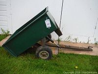 Photo of a green metal garden trailer with the box tilted, resting on grass in front of a white wall. Tires appear flat.