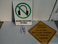 Two signs on white surface: 'Hearing Protectors Optional' plastic sign and yellow diamond-shaped metal comedy sign.