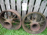 Front view of two antique steel wheels leaning against a wooden fence and standing on grass, showing metal spokes and hollow centers.
