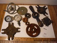 All 12 metal parts shown on a white board with wooden ruler for scale, including gears, pressure gauges, and other mechanical parts.