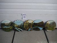 Five circular saw blades with painted folk art landscape scenes arranged on the ground showing variety of nature themes