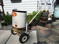 Side-angle view of older CIL lawn spreader showing tub, wheels, handle, and metal frame on stone-paved area.