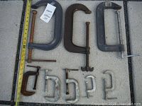 Nine various size 'C' clamps laid out on a grey fabric with a tape measure for scale showing sizes above 8 inches for the large clamps.