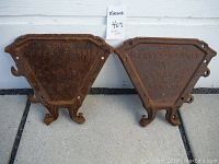 Front side view of two triangular cast iron Massey Harris tractor weights placed upright against a white wall on concrete. Rust clearly visible with embossed branding and model numbers.
