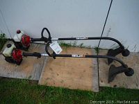 Two Yard Machines string trimmers resting on wooden boards used to hold them upright. Both have red engines and white fuel tanks, black shafts and visible protective guard near the cutting head.