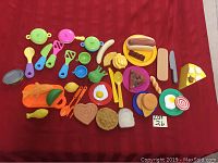 All plastic play food and kitchen accessory items laid out on red cloth.