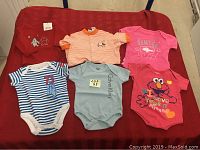 Full lot of 6 baby onesies laid out on red surface showing all items in the lot