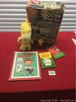 Photo of vintage Electronic Snoopy Playmate in box, Woodstock plush, Charlie Brown figurine, and three Peanuts books displayed on a red cloth.