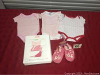 Photo of three single baby onesies, boxed set of five JOE onesies, and a pair of pink baby shoes on a red background