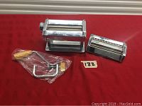 Stainless steel pasta maker set including main roller, cutter attachment, and crank handle laid out on red surface.