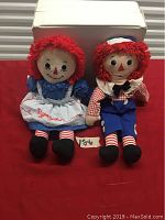 Pair of Raggedy Ann and Andy dolls sitting upright on a red cloth background with a white box behind and a small tag labeled '106'