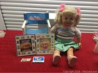 Wide shot of Cricket doll seated next to two books, blue chair with Cricket logo, and cassette tape on table surface.