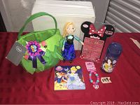 Wide shot of entire lot showing all items together on a red tablecloth, including Tinker Bell green felt basket, Anna plush doll, Minnie Mouse earring sticker display, bracelet, and Belle water bottle.