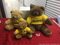 Photo showing three President's Choice teddy bears of different sizes wearing yellow sweaters with their names embroidered, placed on a red cloth background with a white box behind them.