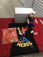 Lot contents with Popeye plush doll, large black t-shirt, small orange t-shirt sealed in plastic, and white box as prop.