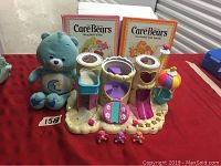 Full lot showing vintage blue Care Bear plush, two vintage Care Bear books, plastic playset, and three small Care Bear figures on red cloth background with lot number tag.