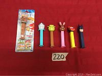 Six Pez dispensers arranged in a line on red background, including Batman, Shrek, Cars character Lightning McQueen, brown bunny, sealed green reindeer, and a blue stem dispenser with white head near the left side.