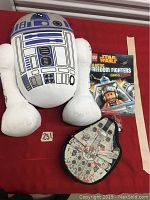 Large R2D2 plush, LEGO Star Wars book, and Millennium Falcon minifigure case all arranged on a red surface.