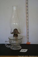 Full view of the entire glass oil lamp with chimney, showing height compared to measuring tape.
