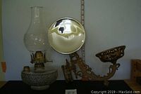 First lamp disassembled showing pressed glass reservoir, metal burner, clear chimney and reflector leaning behind.