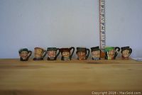 Eight Royal Doulton mini Toby jugs lined up on wooden surface with ruler in background for scale, showing size under 1.5 inches tall.