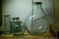 Photo showing five green glass bottles with cork lids arranged on a shelf, sizes range from small to large; visible shapes include round, cylindrical, and square.