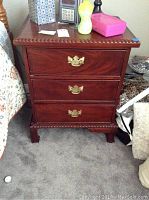 Front view of wood nightstand showing three drawers with gold-tone handles and carved edging