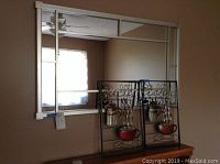 Photo showing fashion wall mirror with segmented frame reflecting room window and ceiling fan; two metal art pieces with scrollwork and small pots placed leaning against mirror.