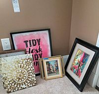 Photo showing various framed art and decor pieces leaning against a wall including pink text print, floral print, and four small framed paintings.