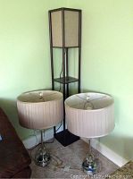 Two ornate table lamps with beige pleated fabric shades showing stains and hanging crystal accents, positioned next to a tall black plastic shelving unit with four shelves.