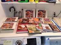 Photo showing assorted cookbooks laid out on a white surface near laundry appliances. Titles include 'Fix It and Forget It Cooking Light for Slow Cookers,' 'Green Juices and Smoothies,' 'Cookies,' and others.