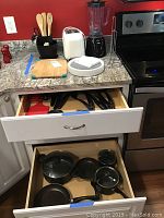 Photo showing kitchen counter with Rival blender, white toaster, bamboo utensils in holder, cutting board, plastic tray and open drawers with pots and pans inside.