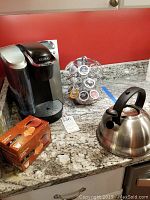 Black Keurig coffee maker, coffee pod carousel with pods, boxed coffee pods, and stainless steel tea pot on granite countertop.