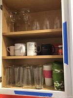 Open cabinet shelf containing glassware and coffee mugs. Top shelf: 4 margarita glasses and 6 stemmed wine glasses. Middle shelf: assorted coffee mugs with various designs including branded and ceramic. Bottom shelf: 12 matching tall drinking glasses with vertical etched lines and 3 travel mugs (green camo and white with pink sleeve).