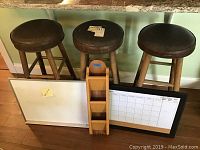Three wooden stools with round black vinyl cushions, showing wear and cracking. Two whiteboards (one plain, one calendar) placed in front. Wooden wall-mounted mail and key holder leaning against the whiteboards.