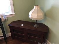 Wooden corner TV cabinet with two glass-paneled doors and a table lamp with metal base and fabric lampshade with fringe, placed on top.