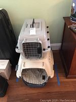 Two stacked pet travel crates against a wall; the top is smaller, the bottom larger, both beige with metal doors.