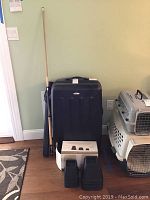 Photo showing black Samsonite hard cover luggage with a wooden cue stick leaning against it, and several black plastic bed risers stacked on the floor, along with two pet carriers to the right.