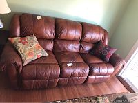 Full view of brown leather recliner couch with two pillows on a wood floor beside a window.