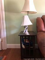 Lamp table with white ceramic accent lamp sitting atop, showing slatted sides and lower shelf with magazines.
