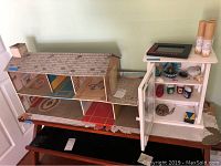 Overall front view of vintage metal dollhouse next to small white cabinet showing various small collectible items inside and on cabinet top.