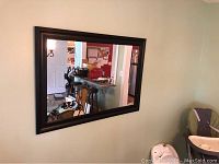 Framed rectangular mirror hanging on a light-colored wall, reflecting part of a room with furniture and entrance door.