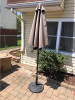 Closed brown outdoor patio umbrella with black round plastic base stand on a patio with pavers in front of a house.