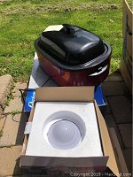 Oster roaster oven with reddish-burgundy base and black high dome lid shown outside with power cord. White ultrasonic humidifier in open box with foam insert visible.