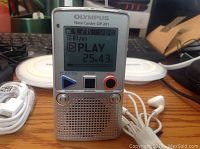 Silver Olympus Note Corder DP-201 digital voice recorder with LCD screen showing playback time, connected white wired earphones, and USB cable