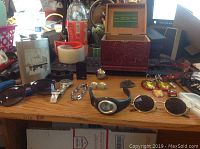 Overview of lot on wooden table showing sunglasses, watches, flask, bracelets, a decorative wooden box, and miscellaneous items in background.